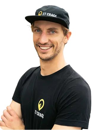 Uśmiechnięty mężczyzna w czarnej czapce z daszkiem i czarnej koszulce, stojący na tle zielonego, naturalnego krajobrazu.